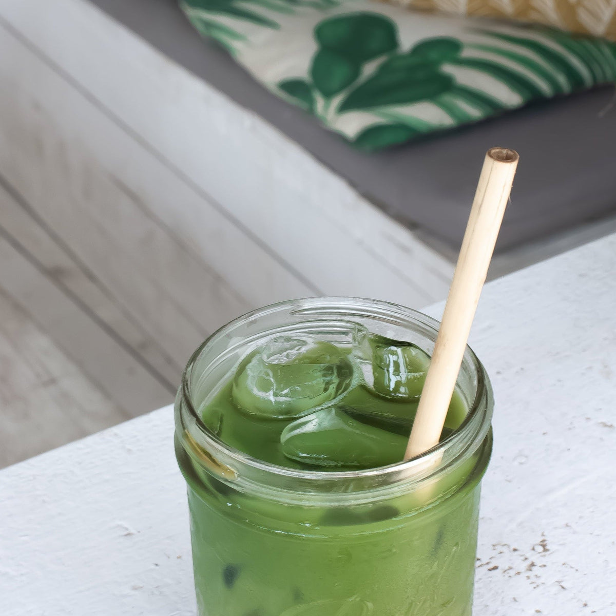Green iced drink in a jar with a straw on a white surface, with a blurred outdoor background.