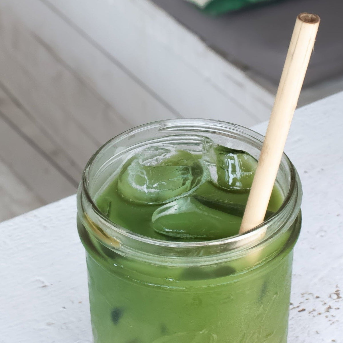 Green iced drink in a jar with a straw on a white surface, with a blurred outdoor background.